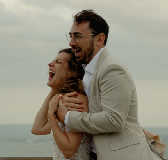 Smiling bride and groom embracing happily outdoors with a cloudy sky background.
