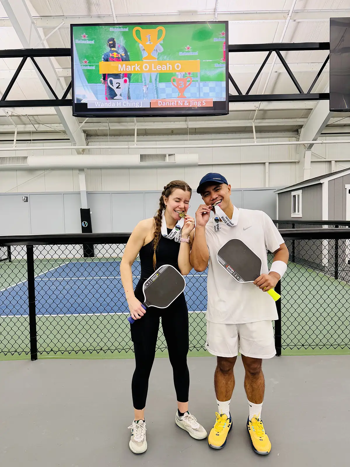Two pickleball players, Mark and Leah Ocampo, bite gold medals while holding paddles on an indoor court.