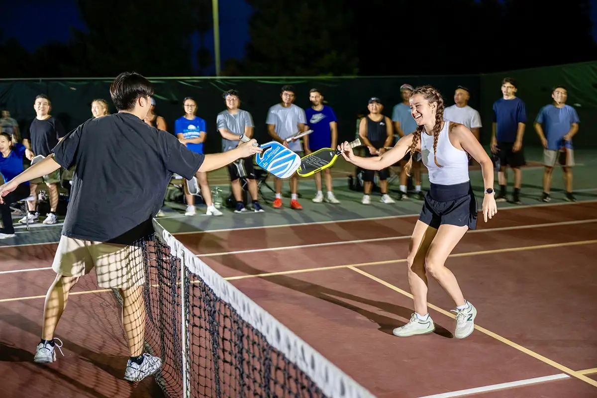 Two players tapping pickleball paddles over the net on an outdoor court at night with a crowd watching behind.