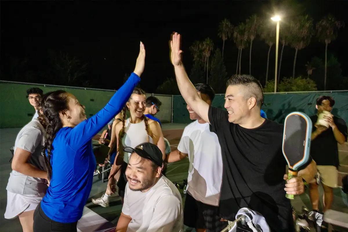 Group of people on an outdoor pickleball court at night, two players giving a high-five, others smiling and socializing.