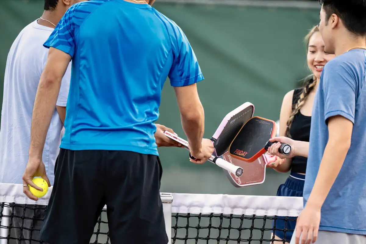 Four people on a pickleball court exchanging paddles over the net, with one person holding a yellow pickleball.