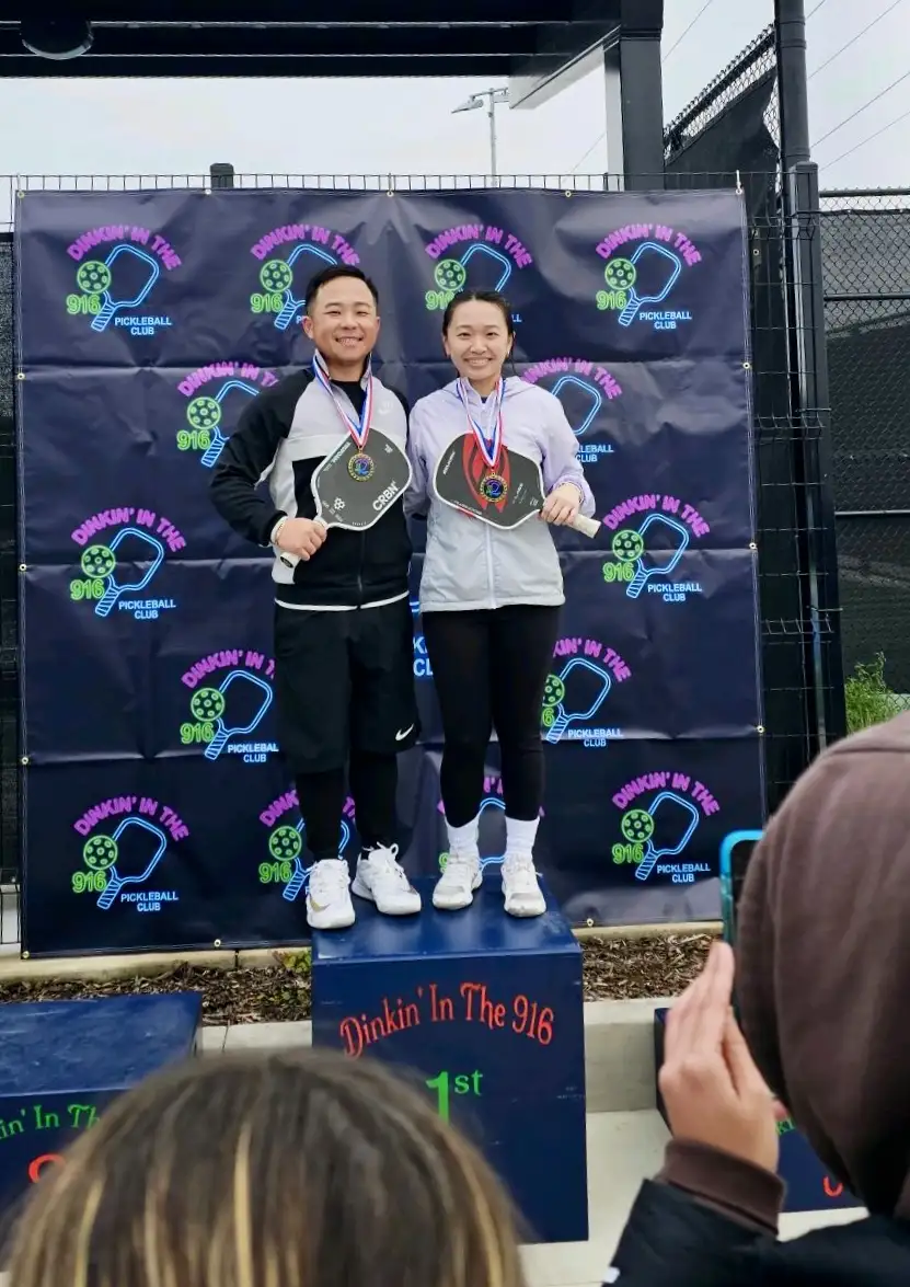 Two pickleball players wearing medals stand on a podium labeled First Place in front of a 'Dinkin' In The 916 Pickleball Club' backdrop.