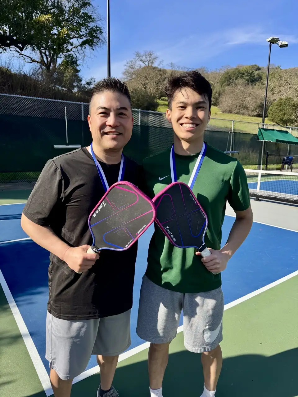 Two men smiling on a pickleball court holding pickleball paddles and wearing medals.