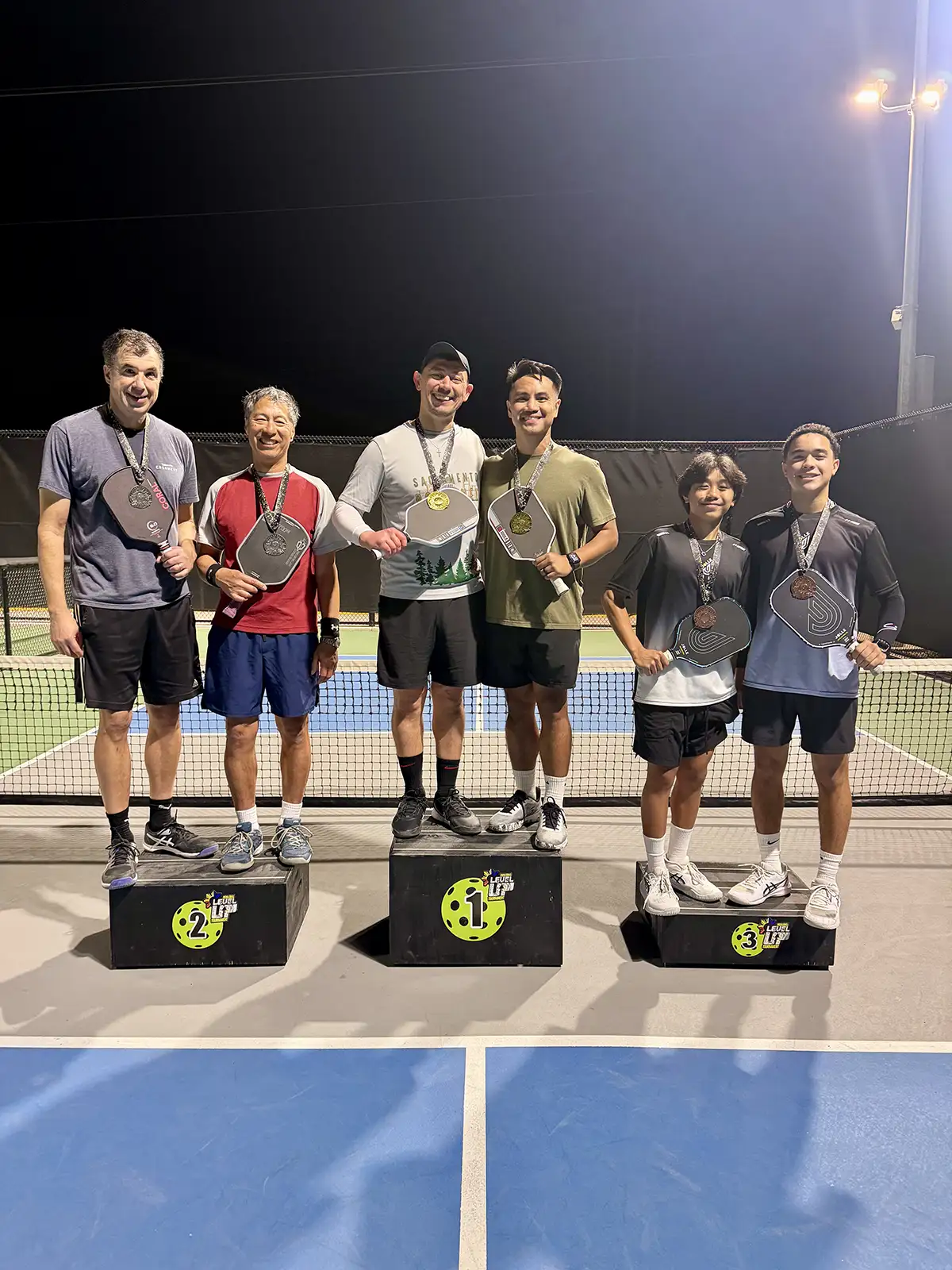 Six men standing on winners podium on a pickleball court at night, each holding paddles and wearing medals.