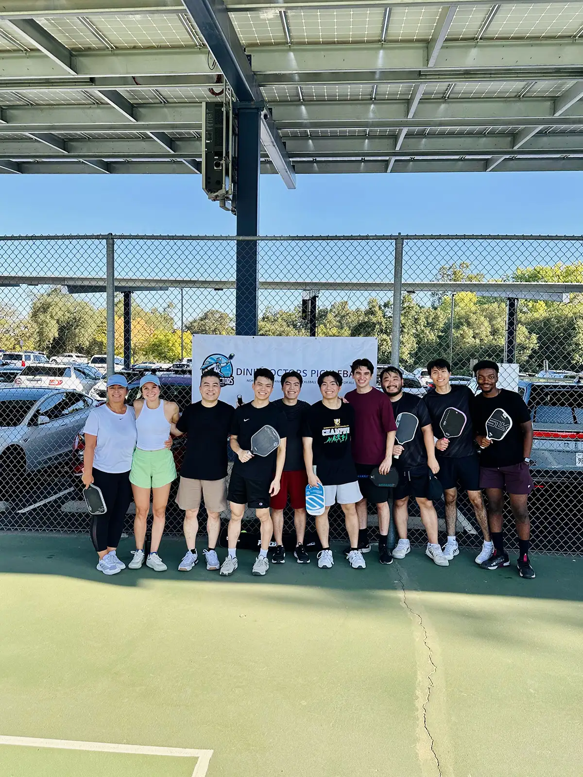 Group of nine people standing on a pickleball court holding paddles, posing in front of a chain-link fence with a banner behind them.