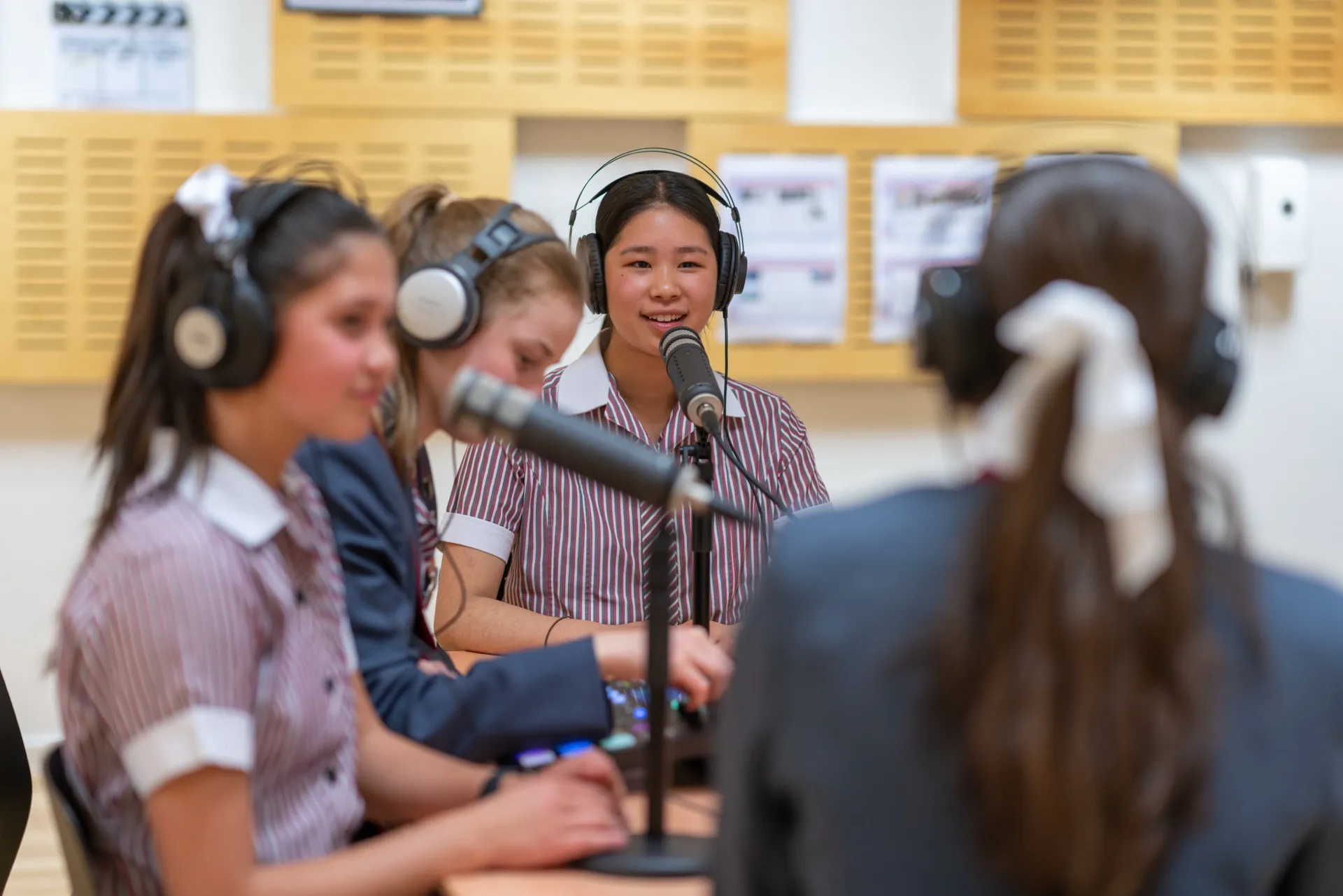 Four teenage girls wearing headphones and striped school uniforms, speaking and recording in a podcast studio.