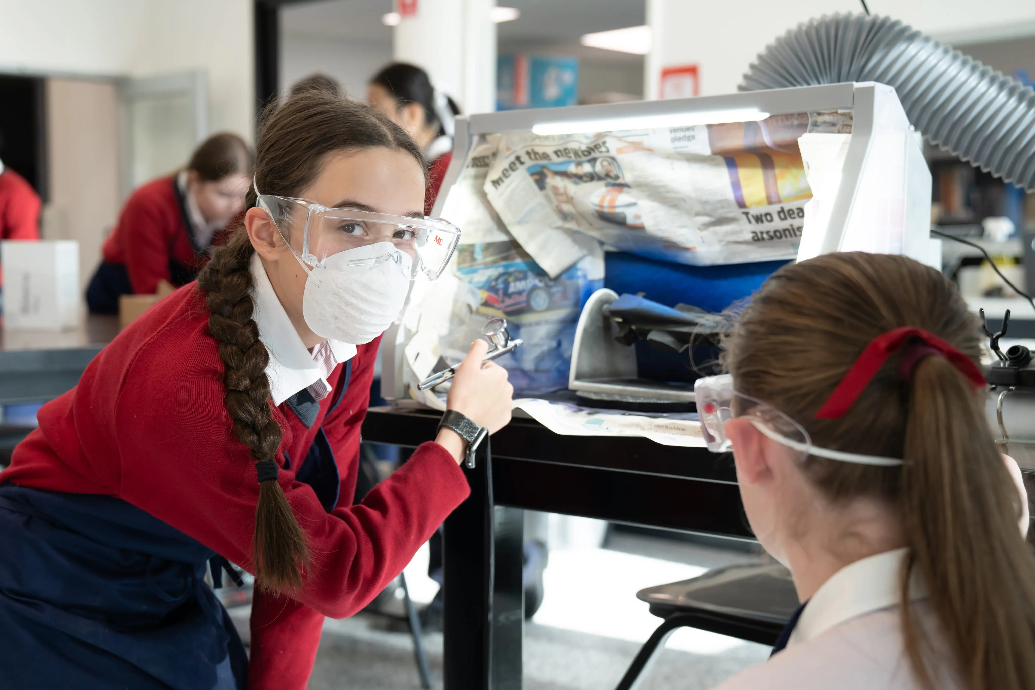 Student wearing protective goggles and mask holds an airbrush while working on a project in a workshop.