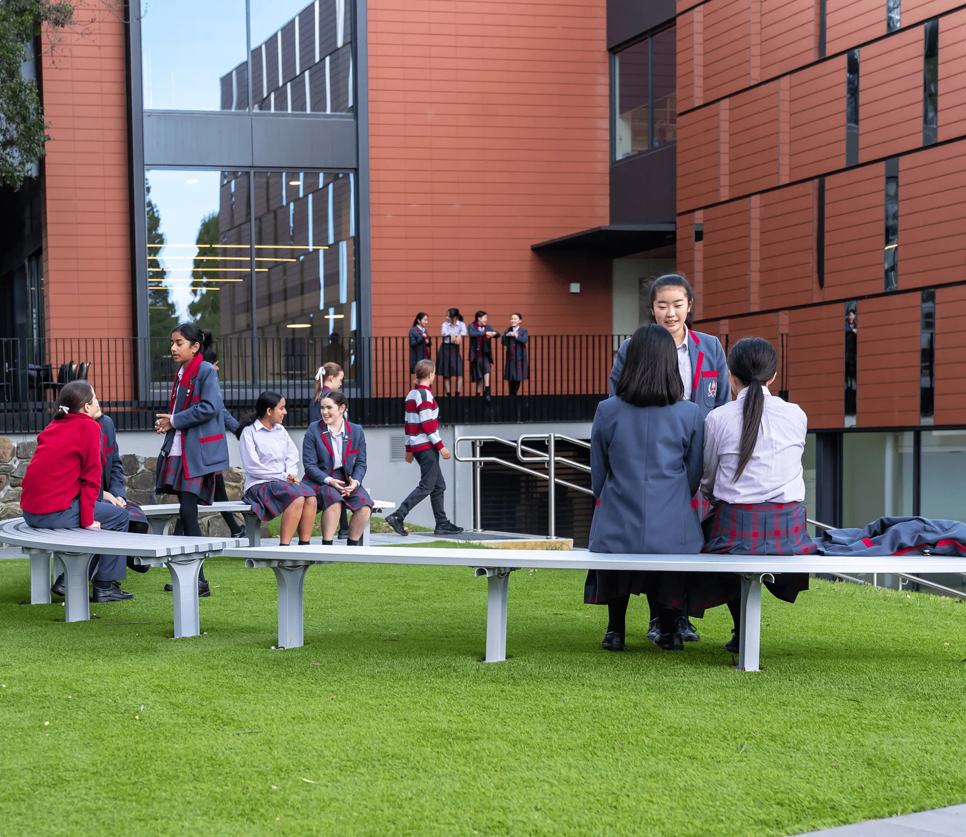 Students in school uniforms socializing on benches on a lawn outside a modern building with orange panels and large windows.