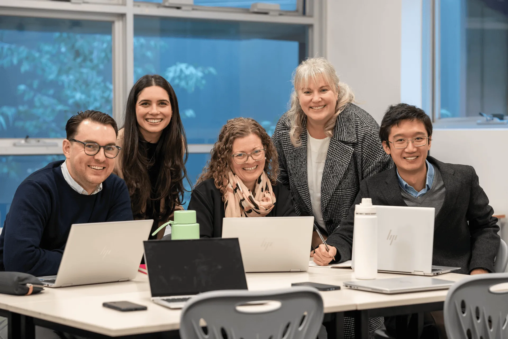 Five diverse professionals smiling and working together at a table with laptops and notebooks in a modern office.