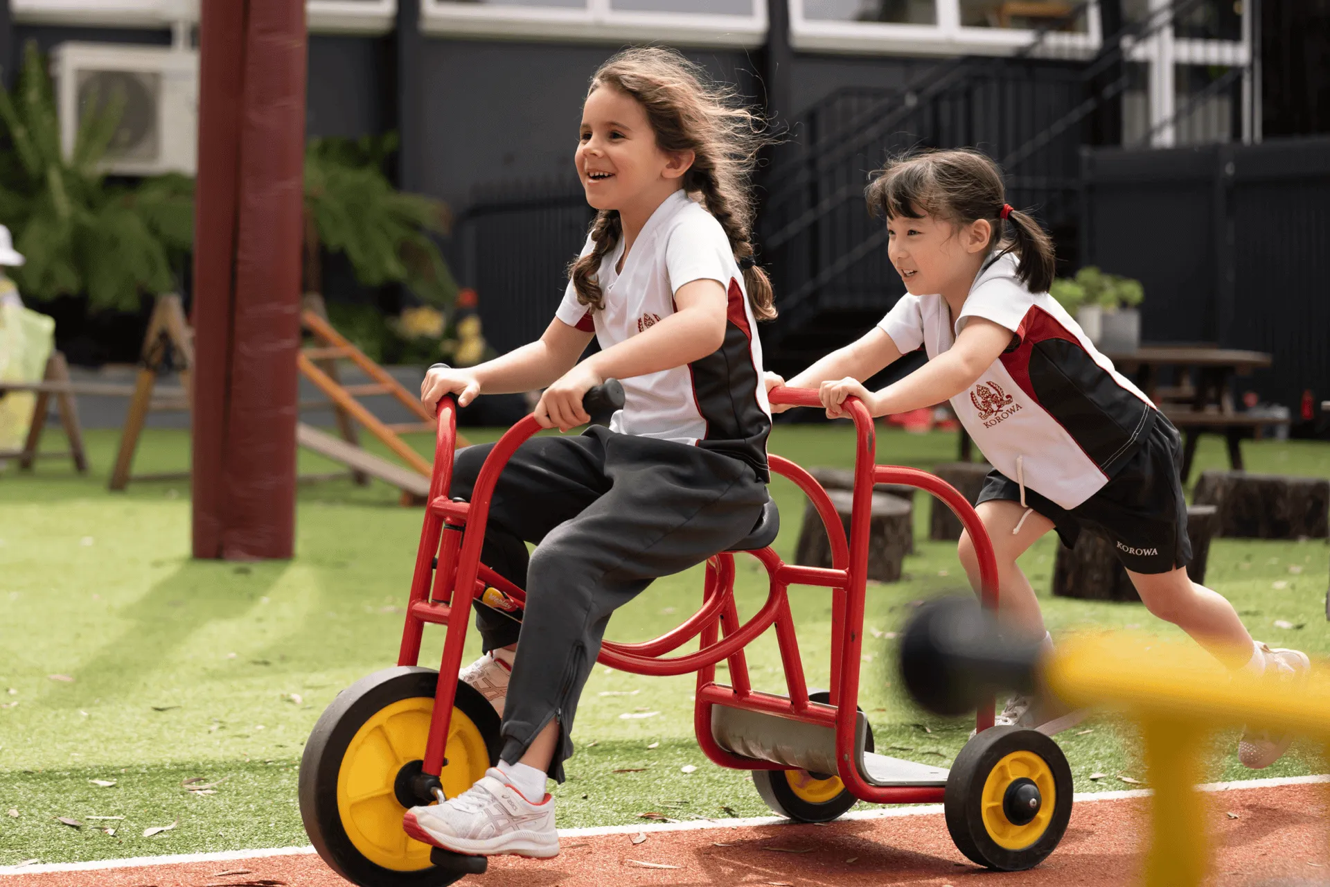 Two girls in school uniforms playing outdoors with a red tricycle, one riding and the other pushing it.