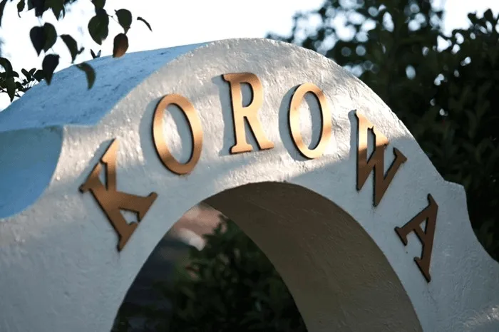 Close-up of a white arched structure with the word 'KOROWA' in large bronze letters on it, surrounded by trees.