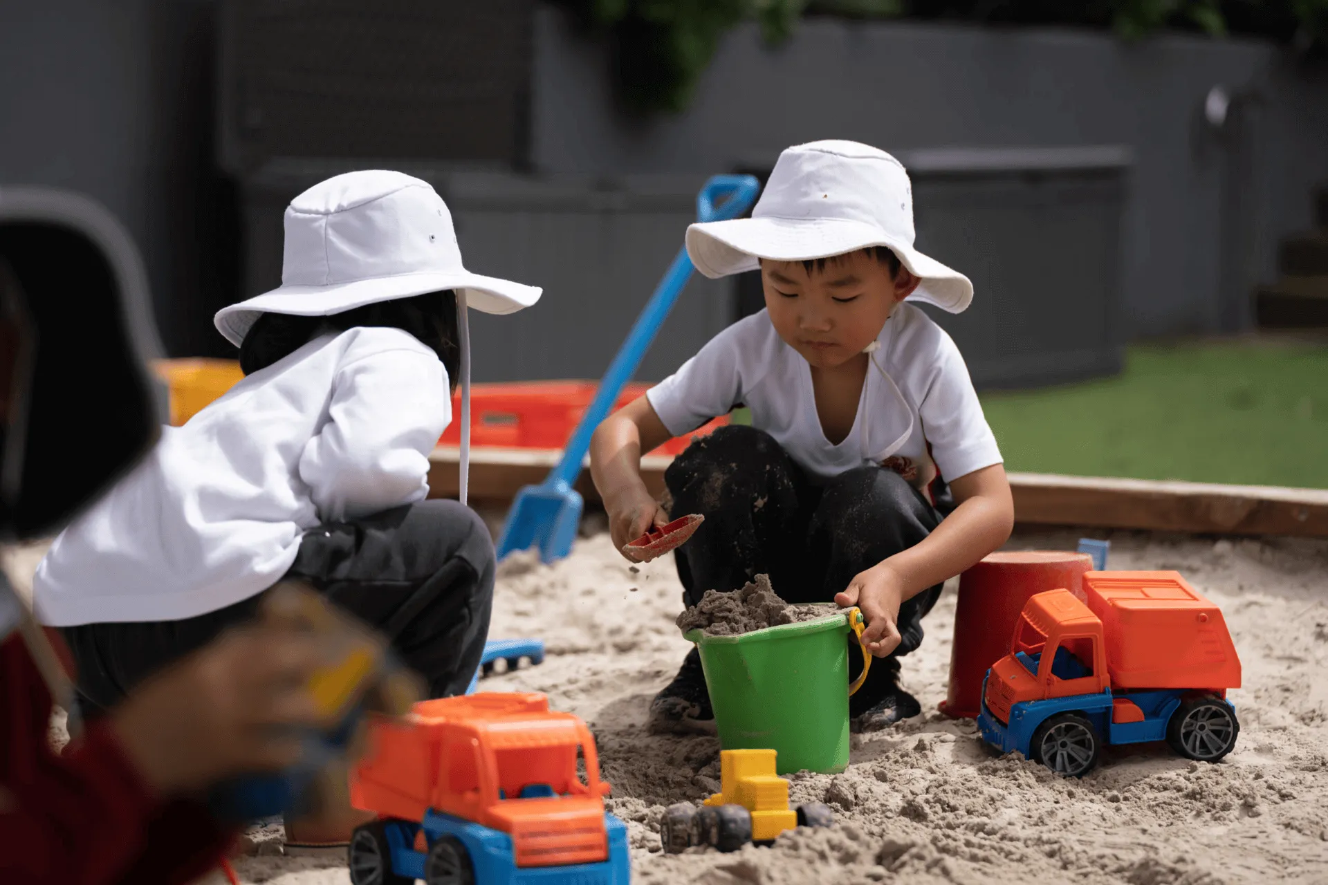 Two children wearing white sun hats playing with sand and toy trucks in a sandbox.