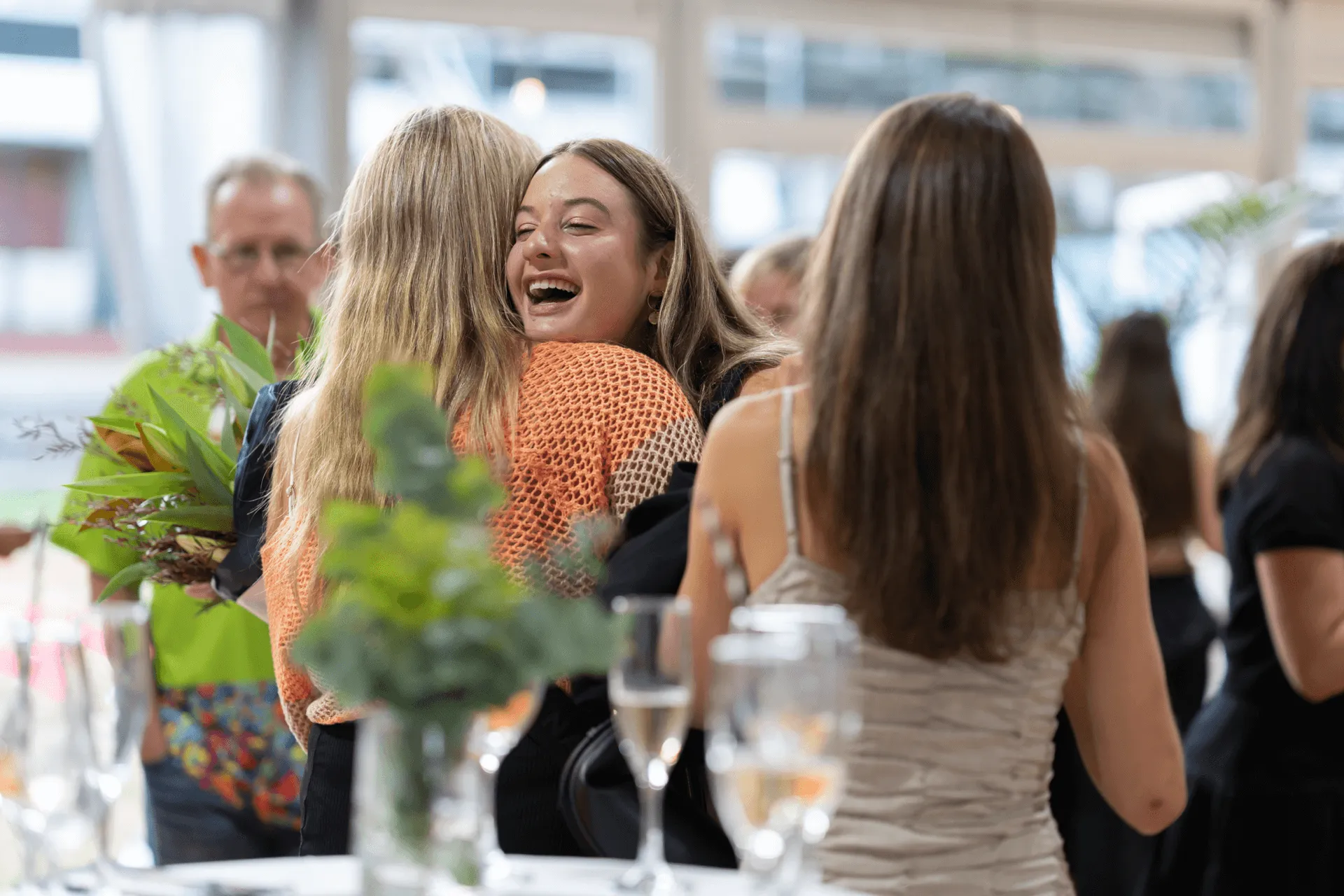 Two women embrace warmly in a lively social gathering with others and drinks in the background.