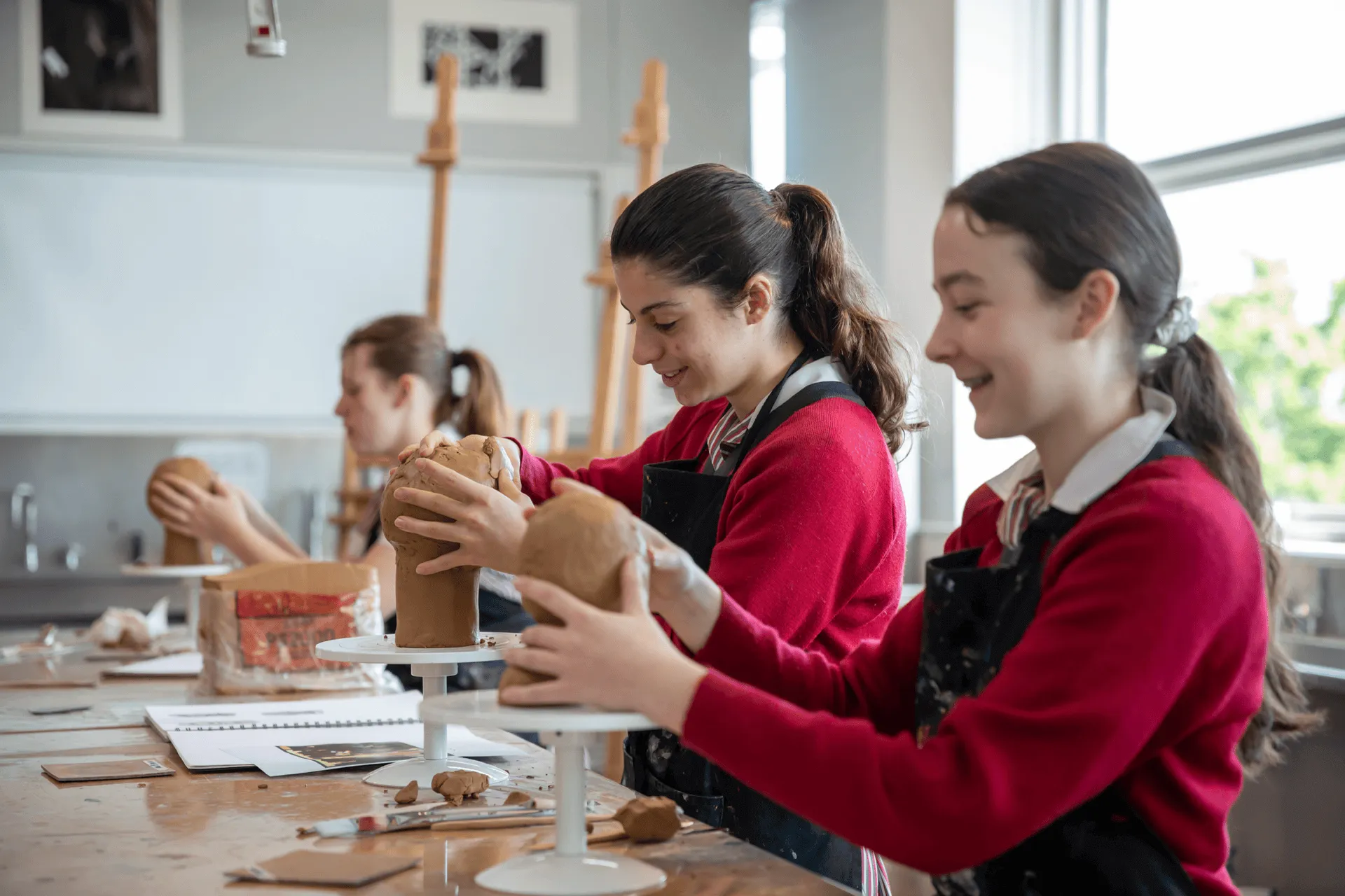Three young women in red sweaters sculpting clay heads in an art classroom.