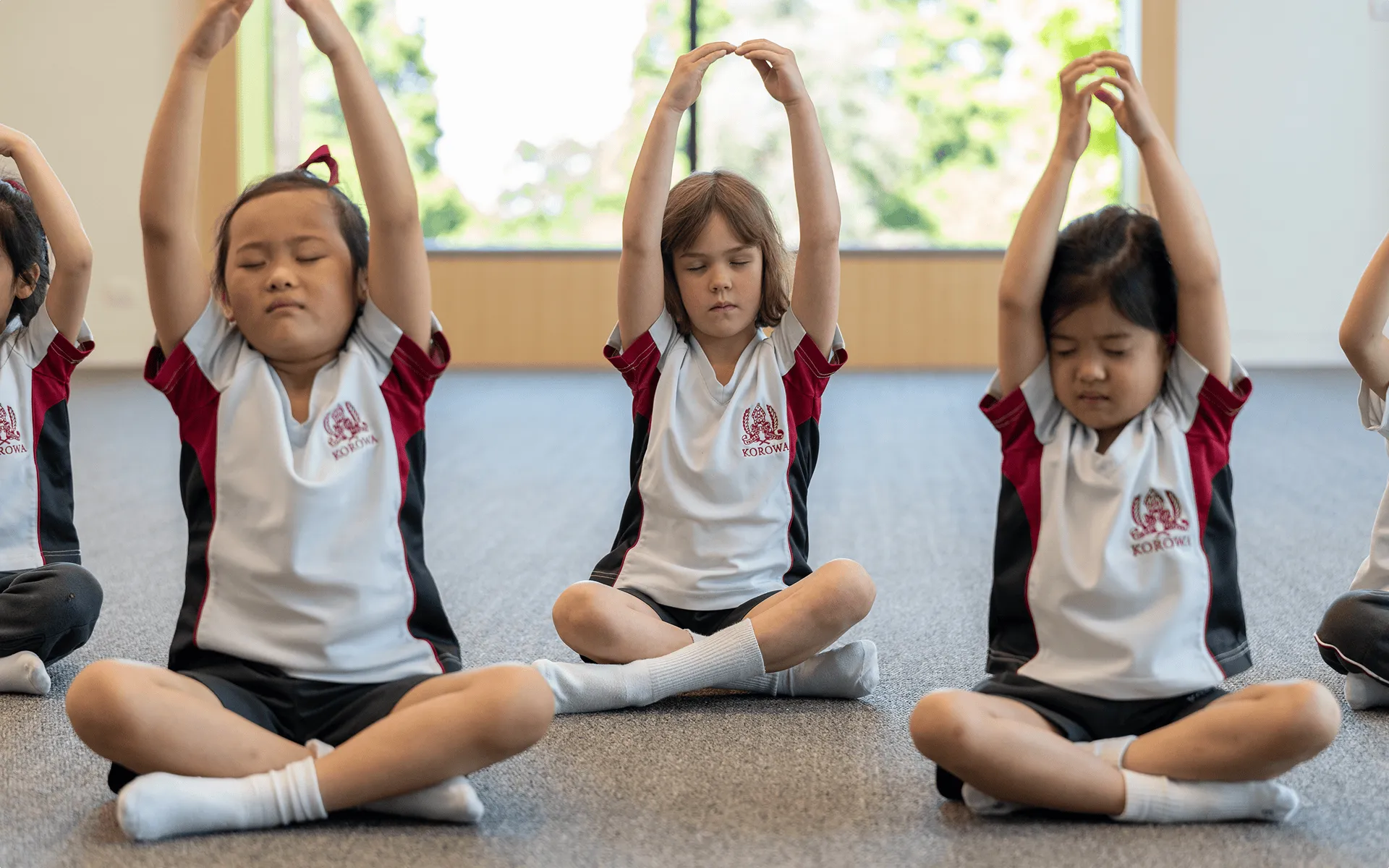 Children sitting cross-legged on a carpet indoors with arms raised above their heads, practicing yoga or meditation.