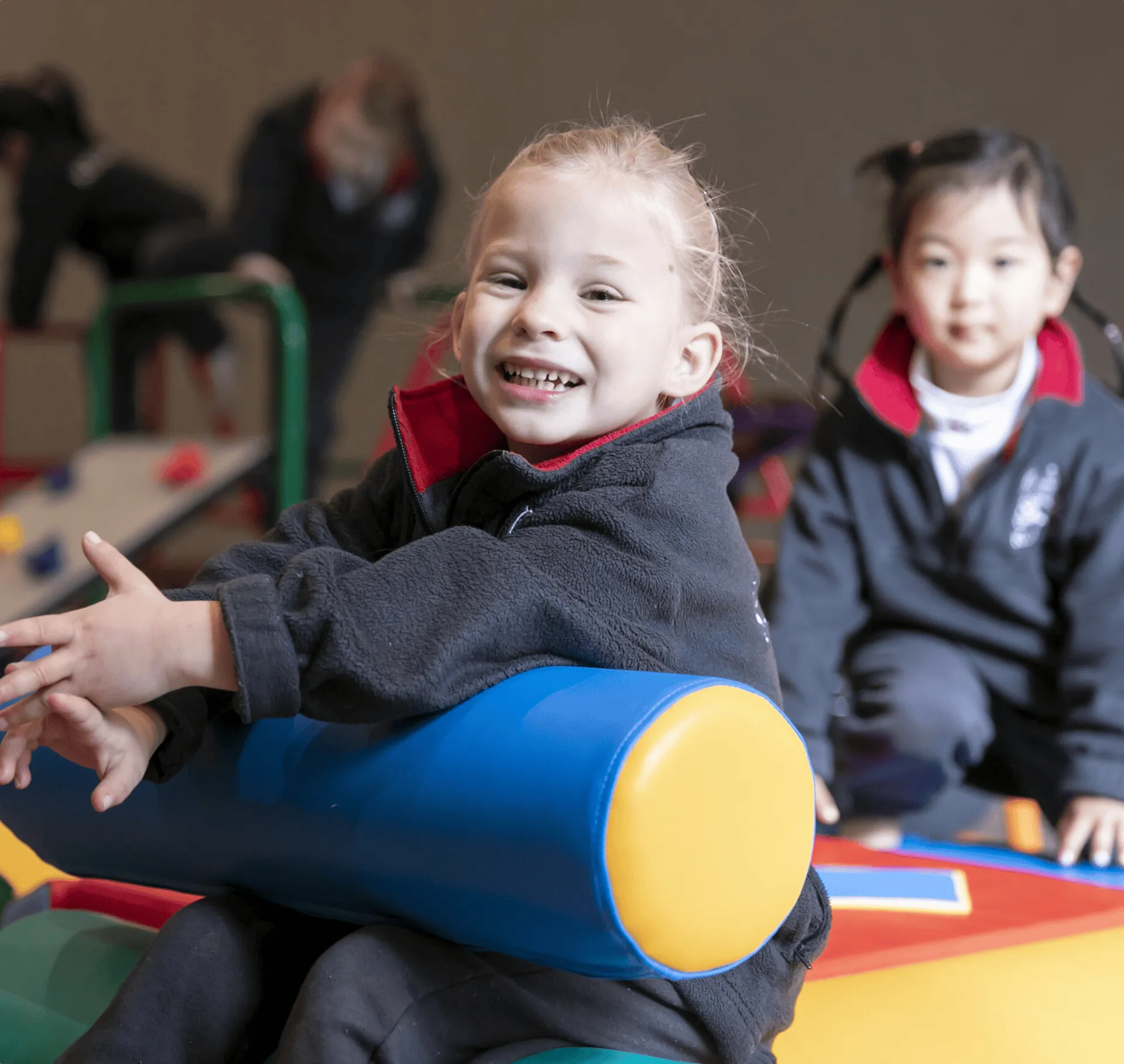 Smiling young girl hugging a blue and yellow cylindrical cushion indoors with another child in the background.