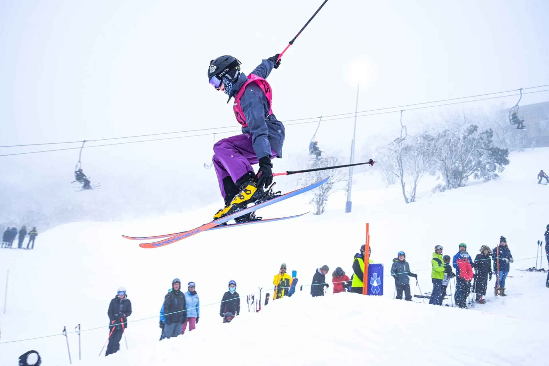 Skier in purple pants and yellow boots airborne performing a jump at a snowy ski resort with spectators and chairlifts in the background.