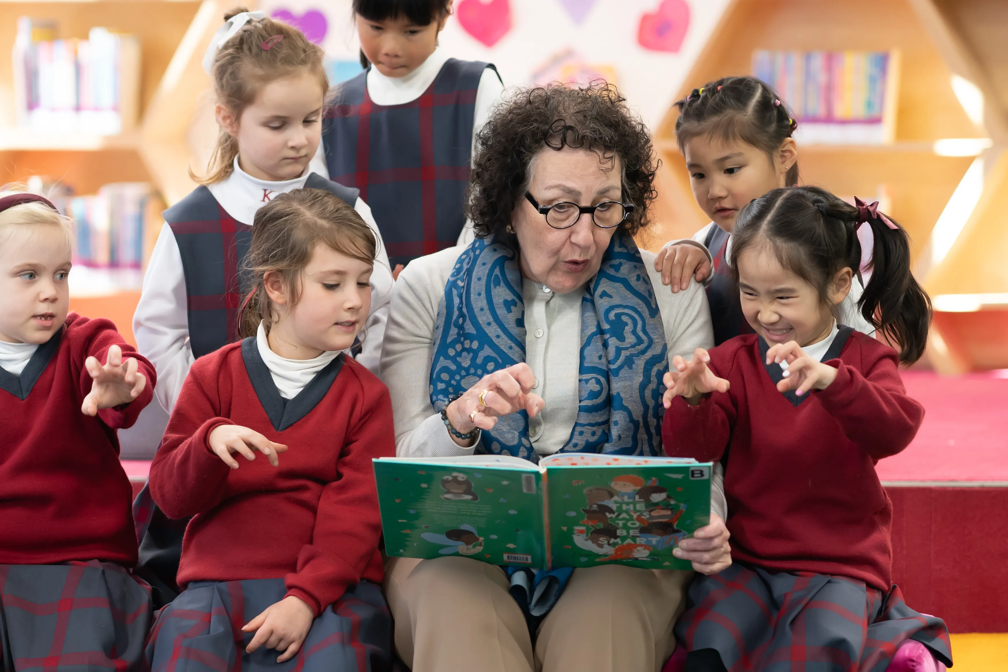 Teacher reading a book titled 'All the Ways to Be Smart' to a group of young girls in school uniforms, with the children making playful claw gestures.