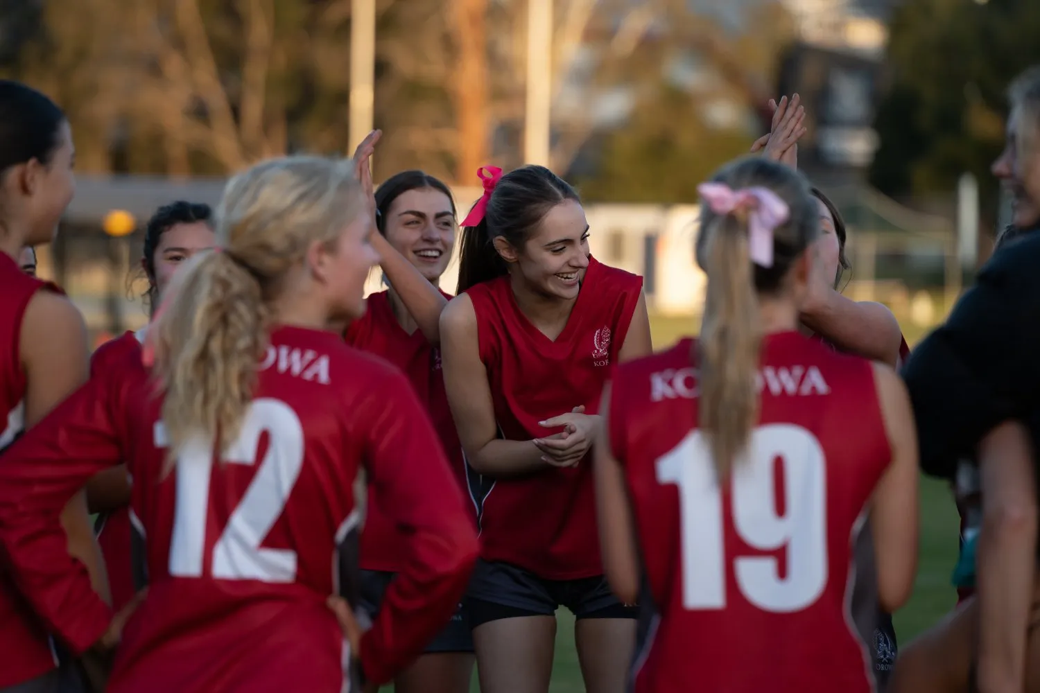 Female athletes in red jerseys numbered 12 and 19 celebrating and smiling outdoors on a sports field.