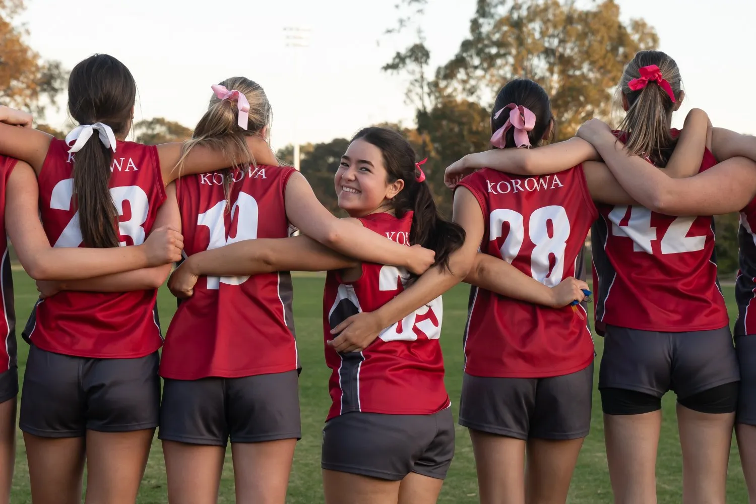 Girls' sports team in red and black uniforms standing arm in arm, one girl smiling at the camera.