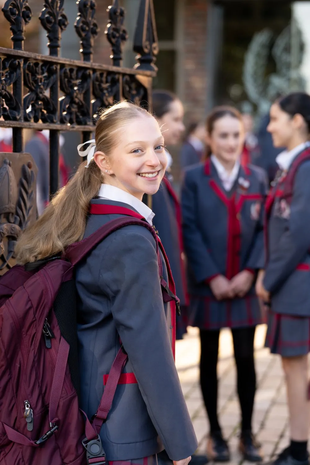 Smiling schoolgirl with a maroon backpack standing near a wrought iron fence, with three other students in uniform blurred in the background.