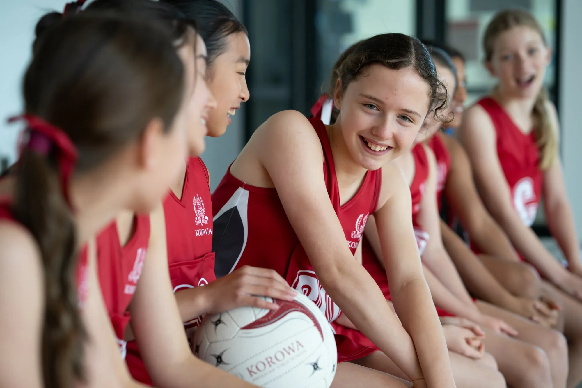 Smiling girls in red Korowa sports uniforms sitting on a bench, holding a netball.