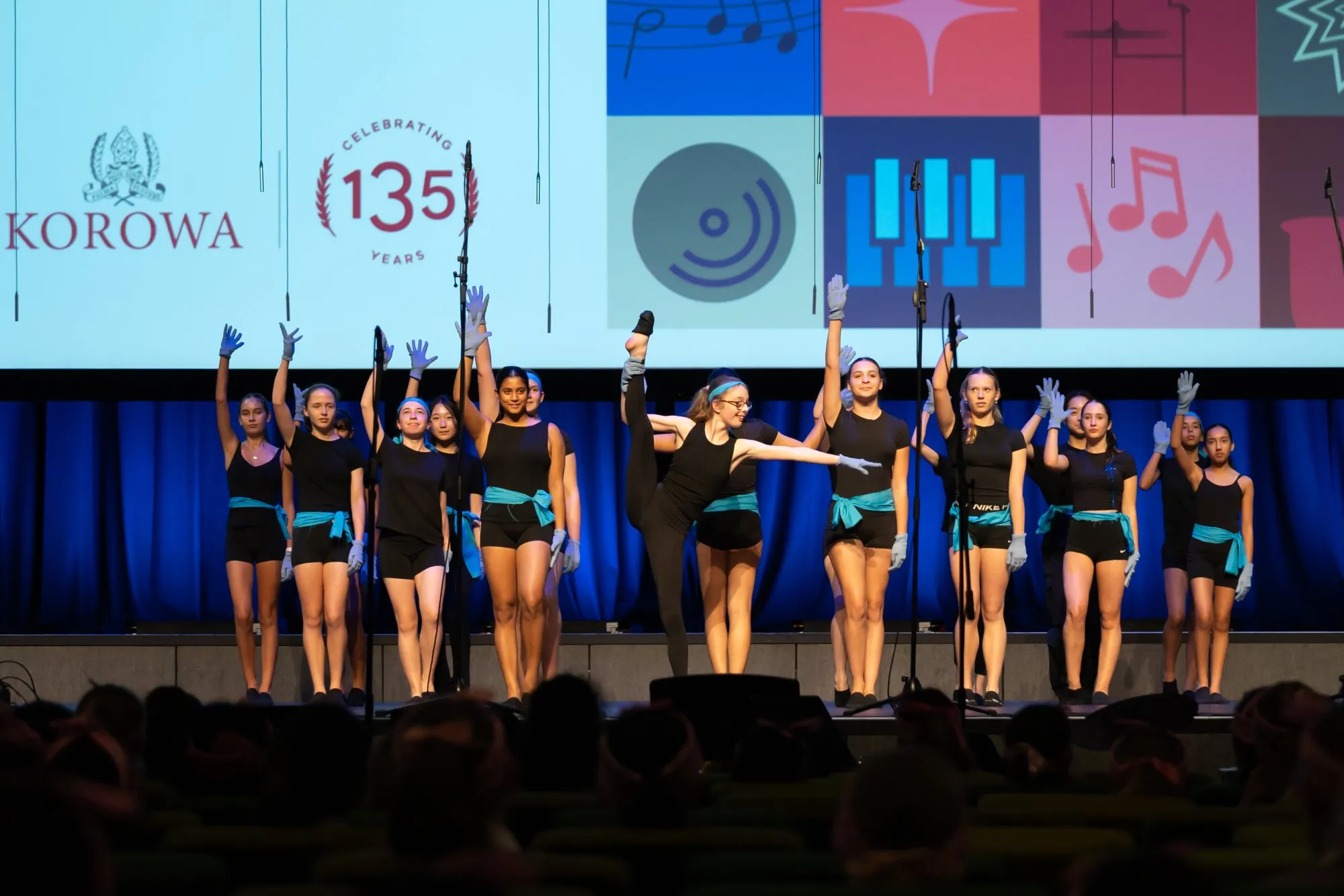 A group of young dancers wearing black outfits with blue sashes and gloves performing on stage, one dancer in the center stretching her leg upward.
