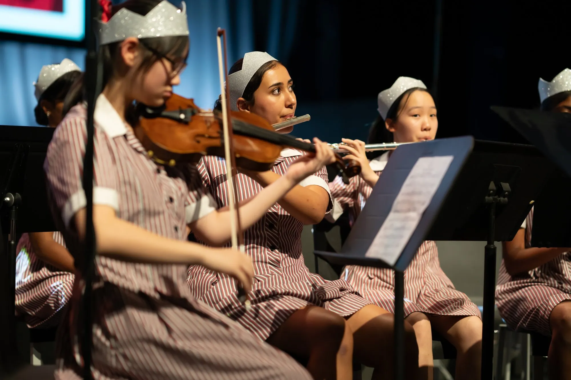 Young female students in striped uniforms and silver crowns playing violin and flute in a school orchestra.