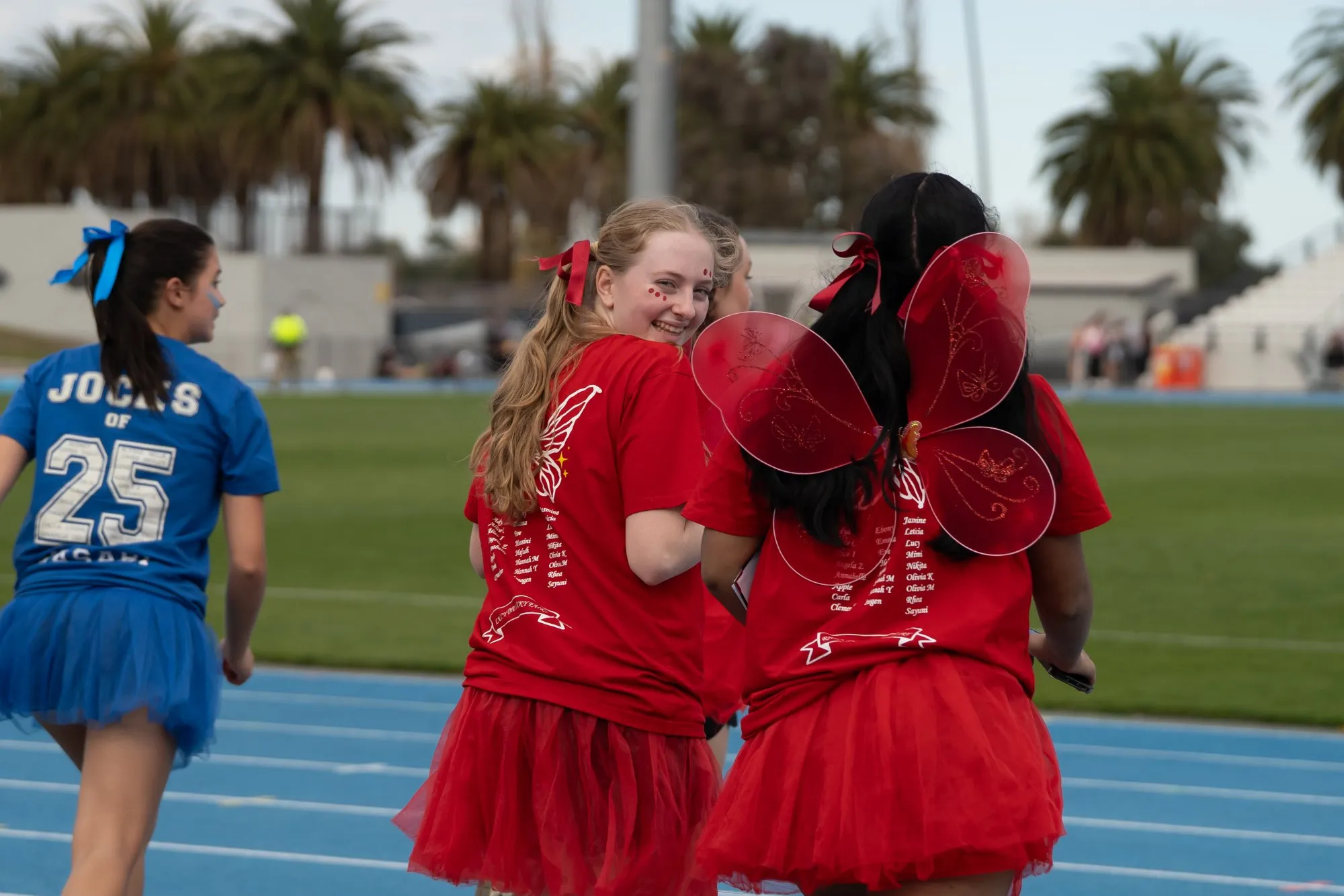 Two girls in red tutus and shirts, one wearing red butterfly wings, smiling and walking on a blue running track with a girl in a blue tutu in the background.
