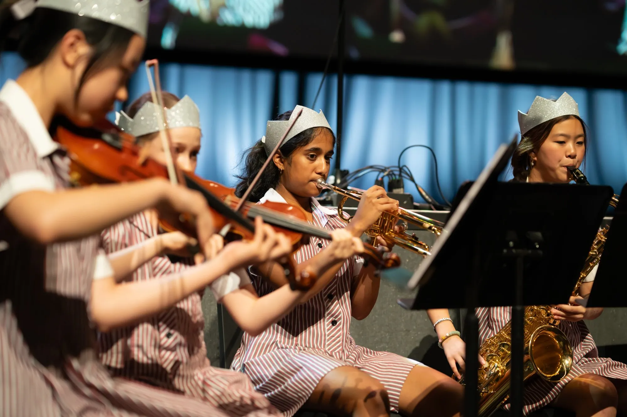 Four young female musicians wearing striped uniforms and silver crowns playing violin, trumpet, and saxophone in a band performance.
