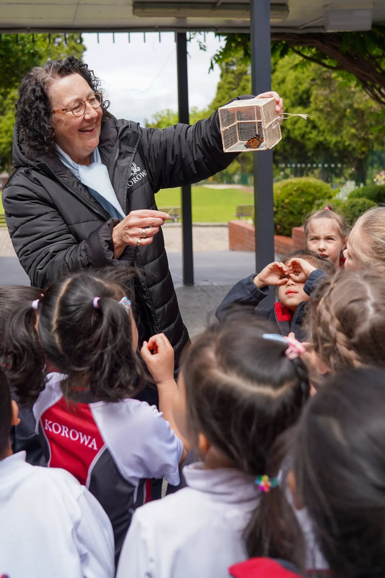Smiling woman holding a cage with a butterfly, surrounded by children watching attentively outdoors.