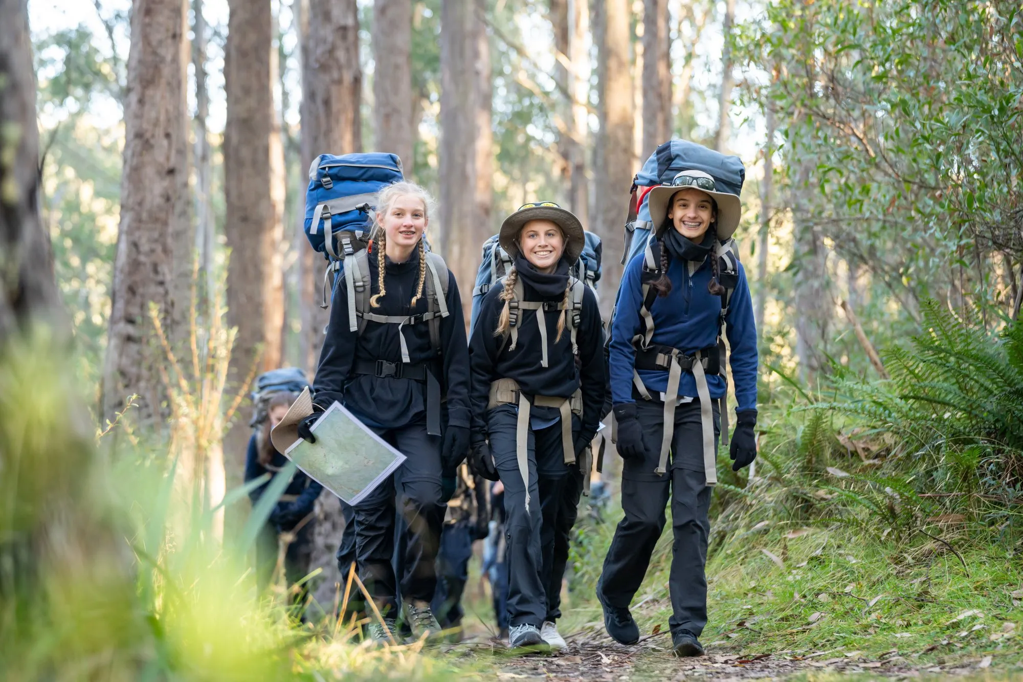 Three teenage girls hiking on a forest trail wearing backpacks and outdoor gear, one holding a map.