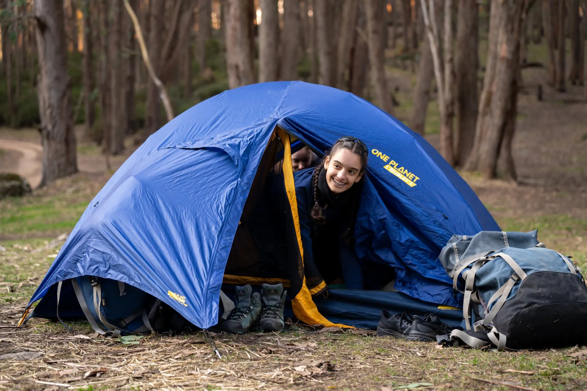 Smiling girl with braided hair peeking out from a blue camping tent in a forest, with hiking boots and backpacks nearby.