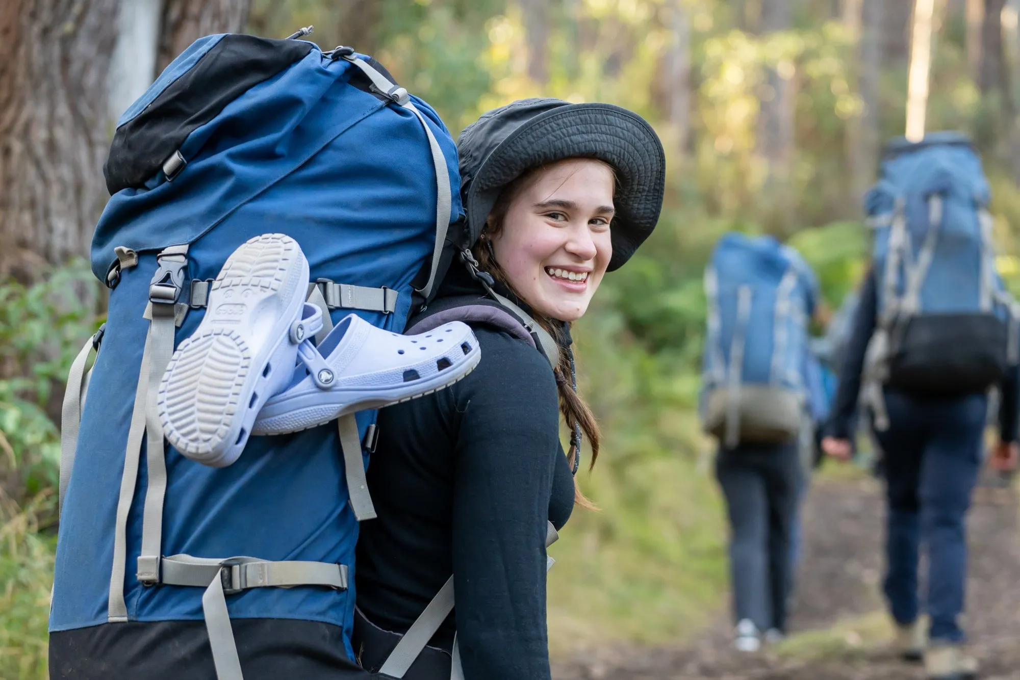 Smiling young woman wearing a black hat and carrying a large blue backpack with white shoes attached, hiking with two others on a forest trail.