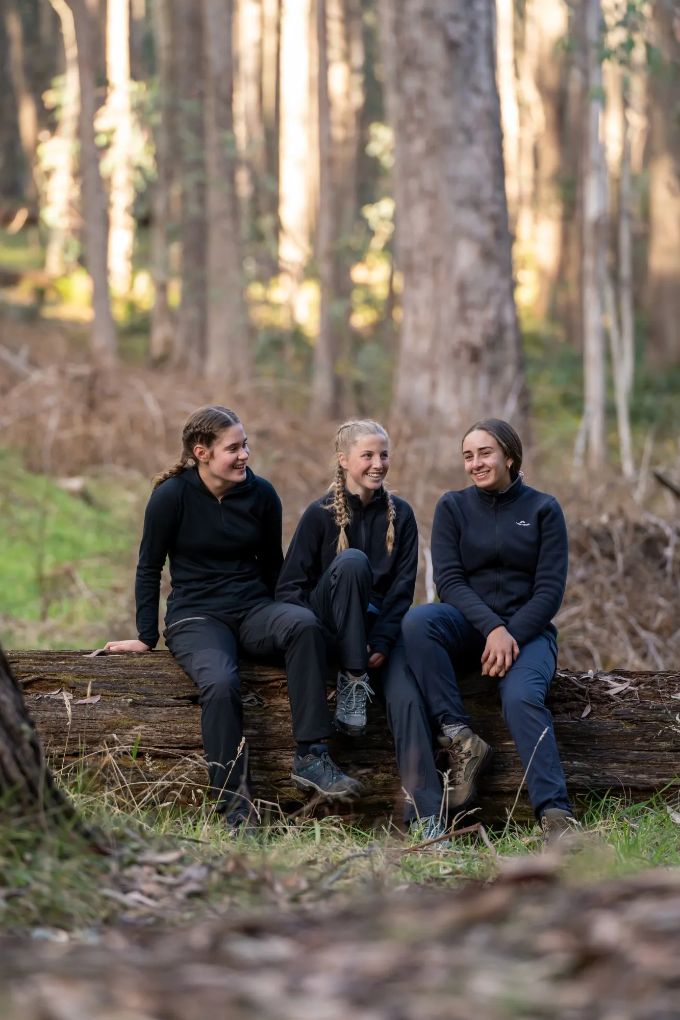 Three young women sitting on a fallen tree trunk in a forest, smiling and wearing outdoor hiking clothes.