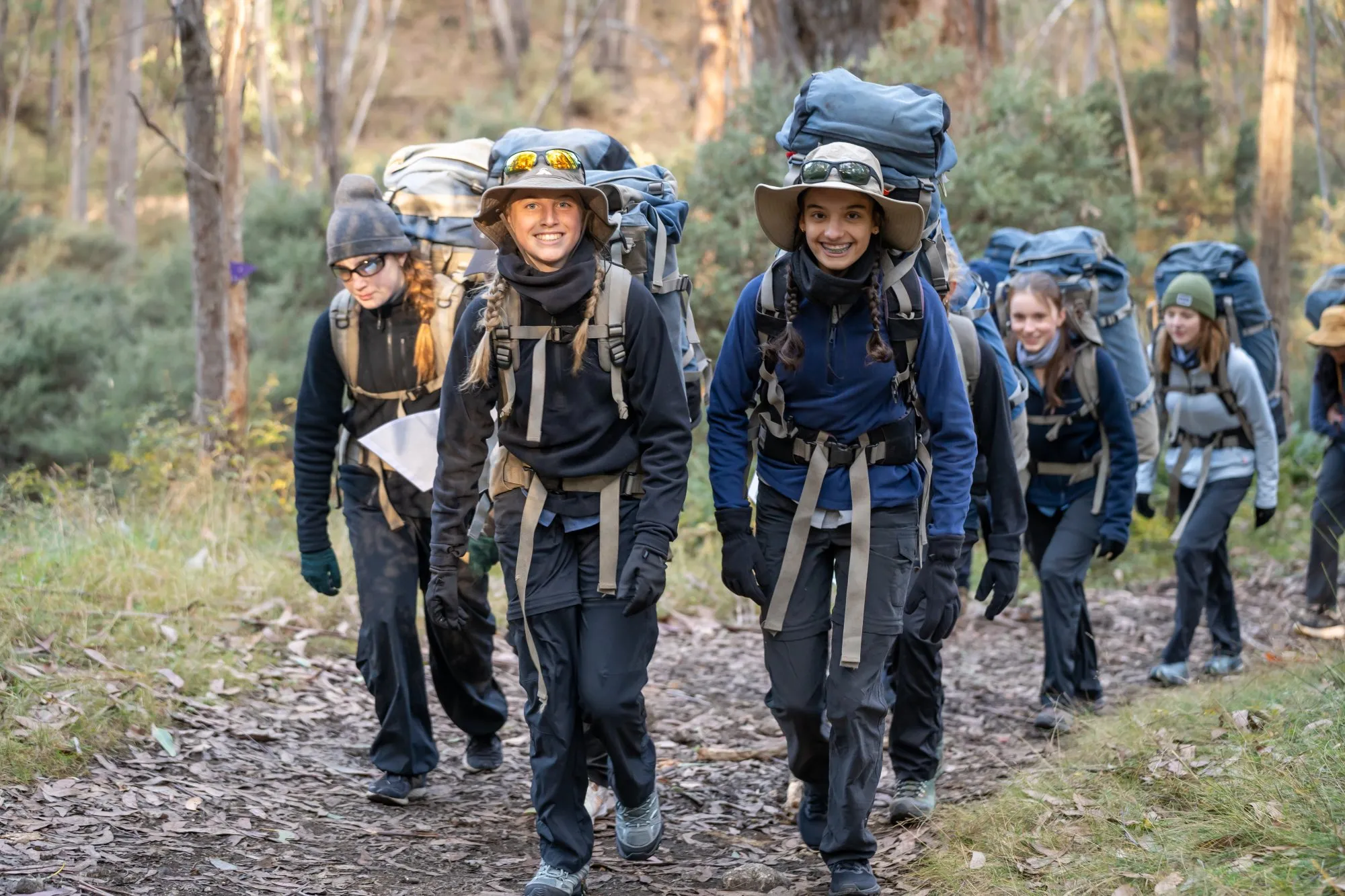 Group of smiling young hikers carrying large backpacks walking on a forest trail.