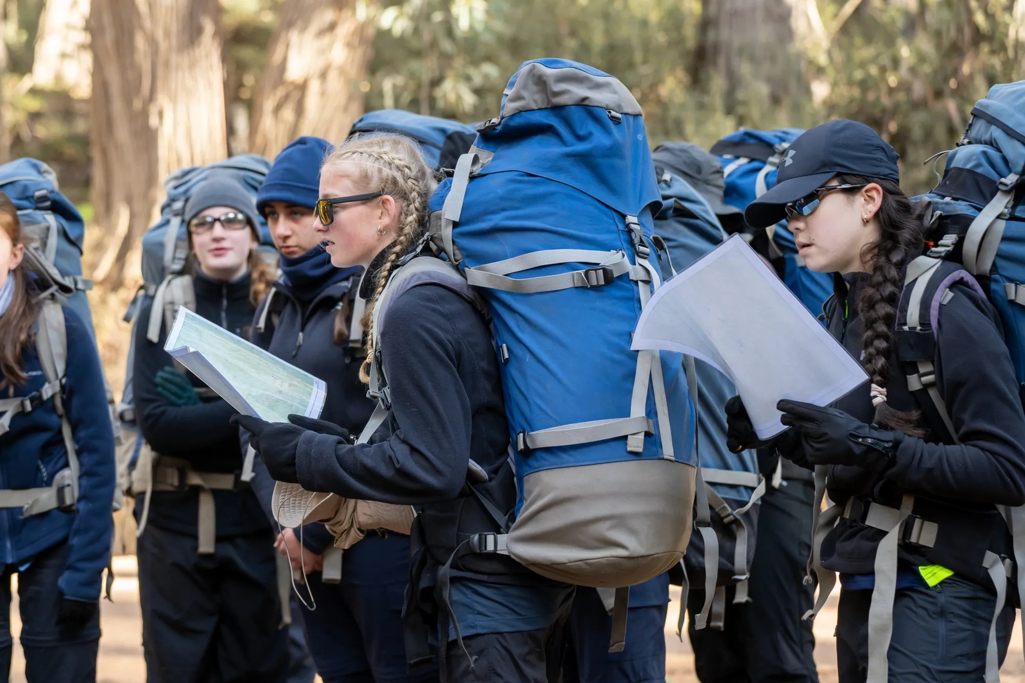 Group of young hikers with large blue backpacks examining maps in a forest setting.