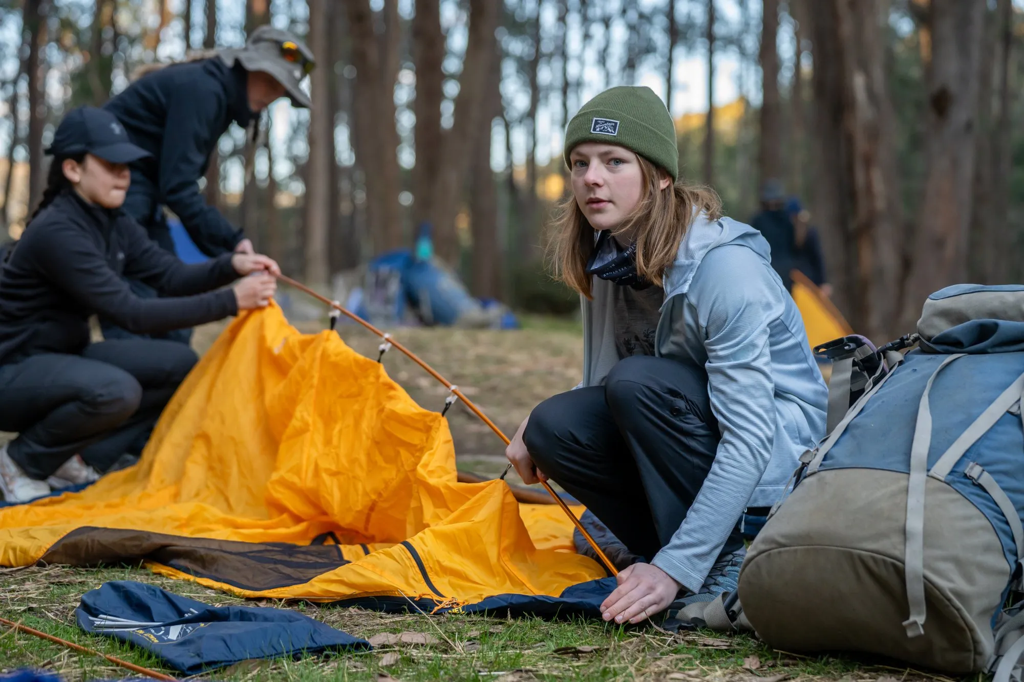 Three people setting up an orange tent in a forest campsite with backpacks nearby.