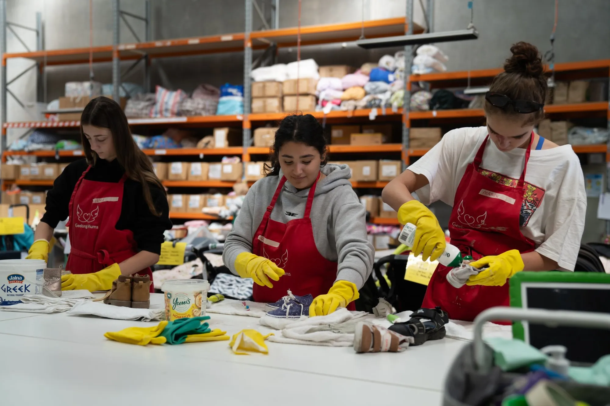 Three women wearing red aprons and yellow gloves cleaning and organizing small shoes and items on a white table in a warehouse.