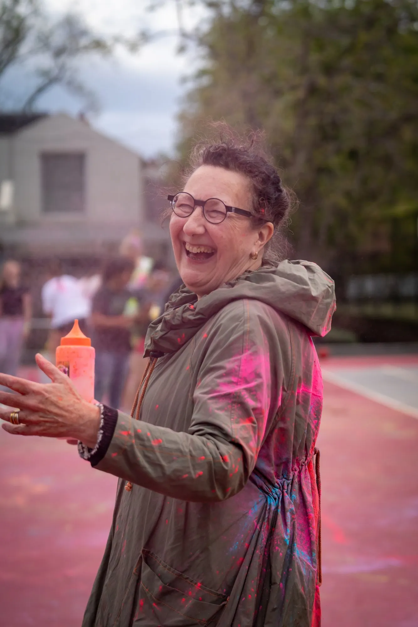 Smiling woman wearing glasses and a green jacket with colorful paint splatters, holding an orange squeeze bottle outdoors.