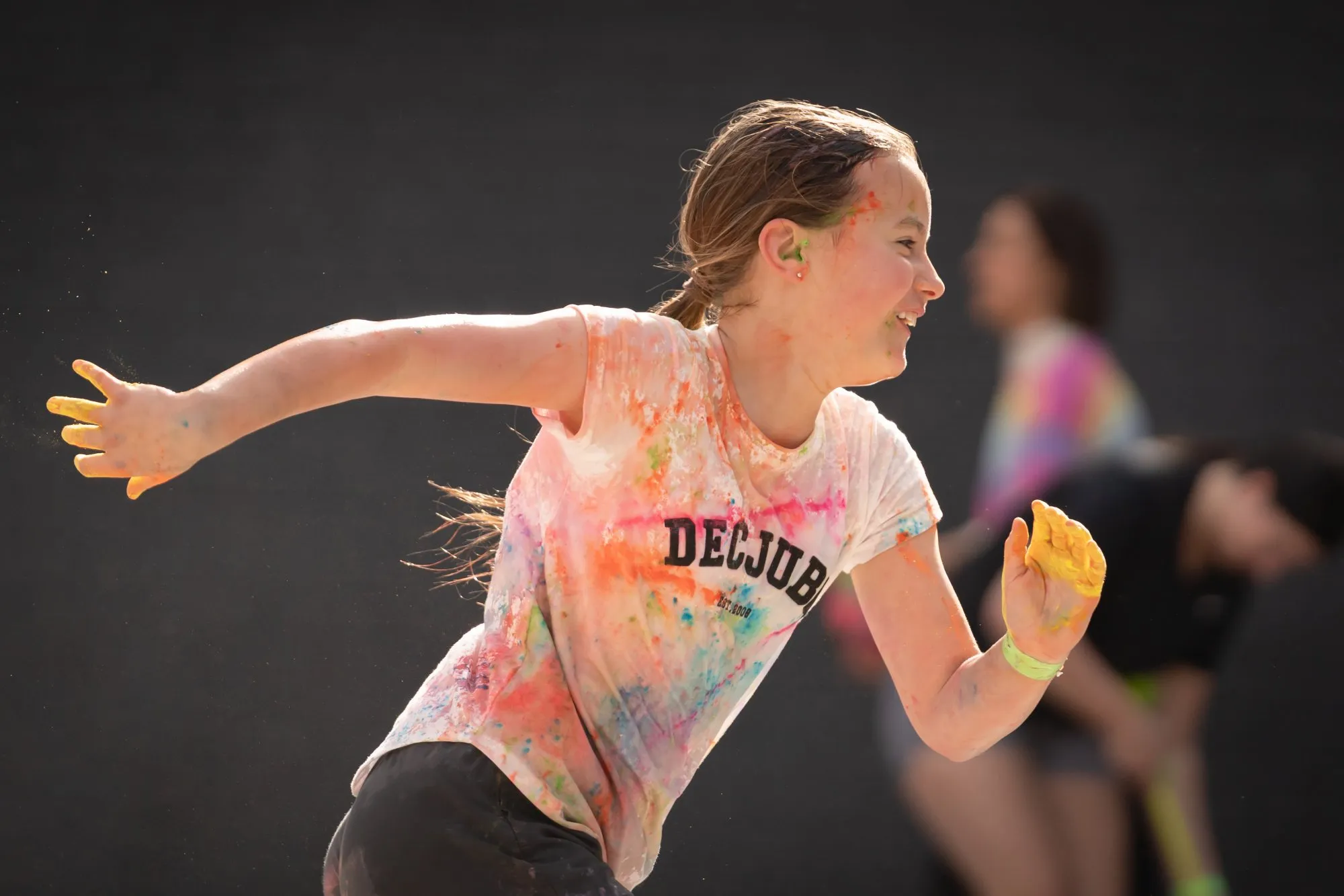 Smiling girl with colorful paint stains running outdoors with blurred people in the background.