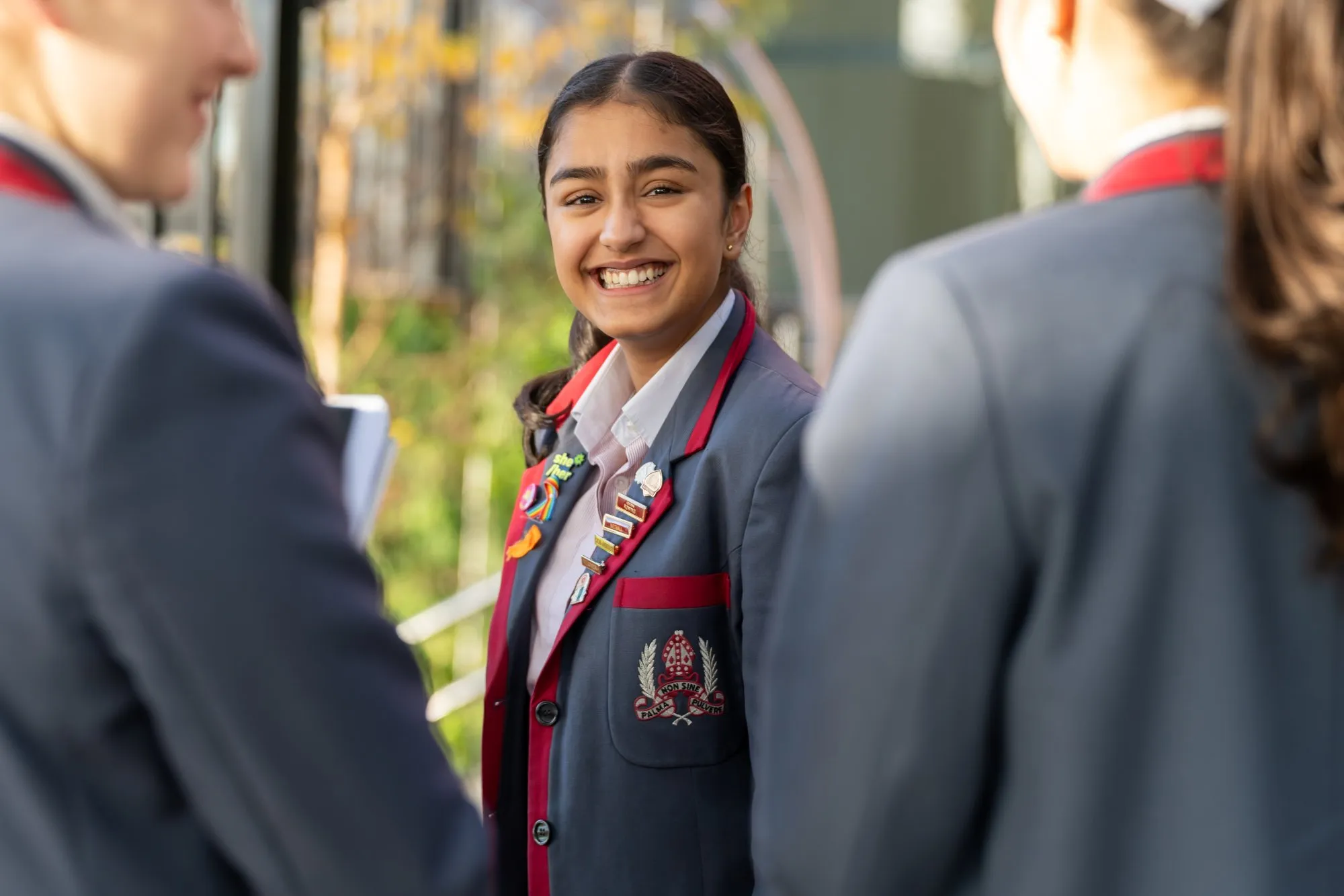 Smiling teenage girl in a navy blazer with red trim and badge standing outdoors between two other students.