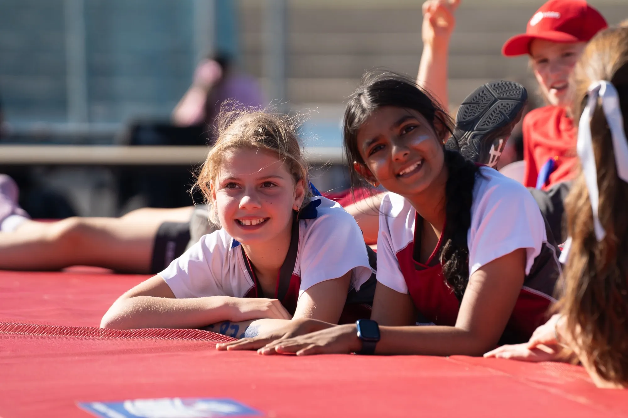 Two girls smiling and lying on a red mat outdoors during a sports event.