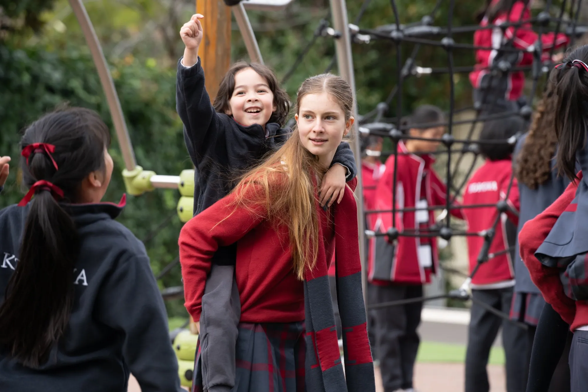 Smiling girl giving a piggyback ride to a laughing child pointing upward in a playground, with other children and climbing equipment in the background.