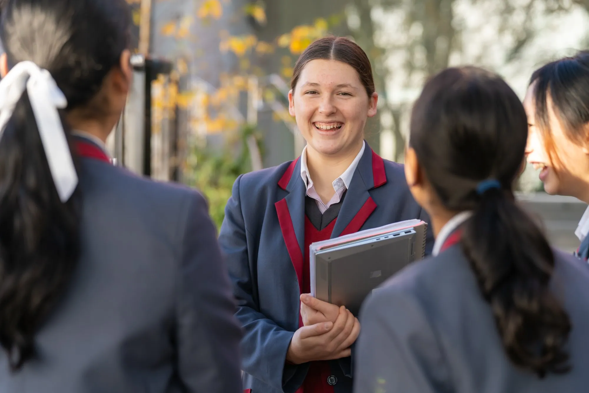Smiling student in a navy and burgundy school uniform holding notebooks, talking with three other students outdoors.