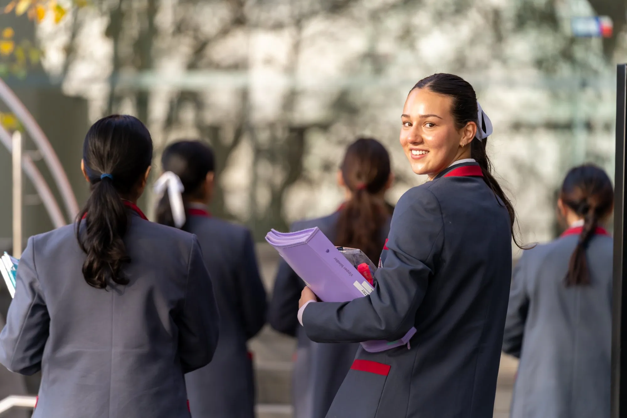 Smiling female student in a school uniform holding a purple folder, looking back while walking with other students in the background.