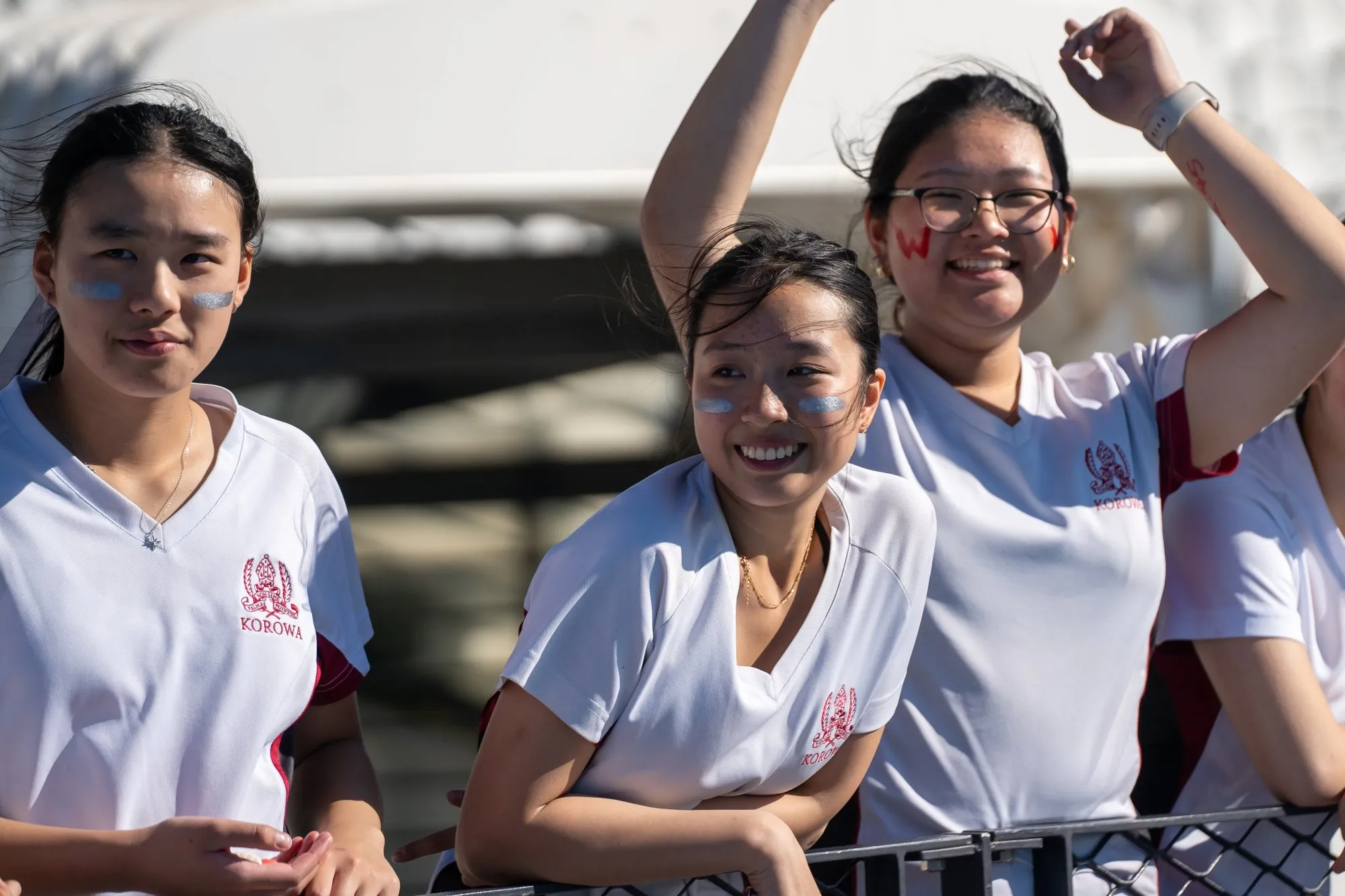 Three young women wearing white Korowa sports shirts, two with blue face paint and one with red face paint, smiling and standing behind a fence in sunlight.