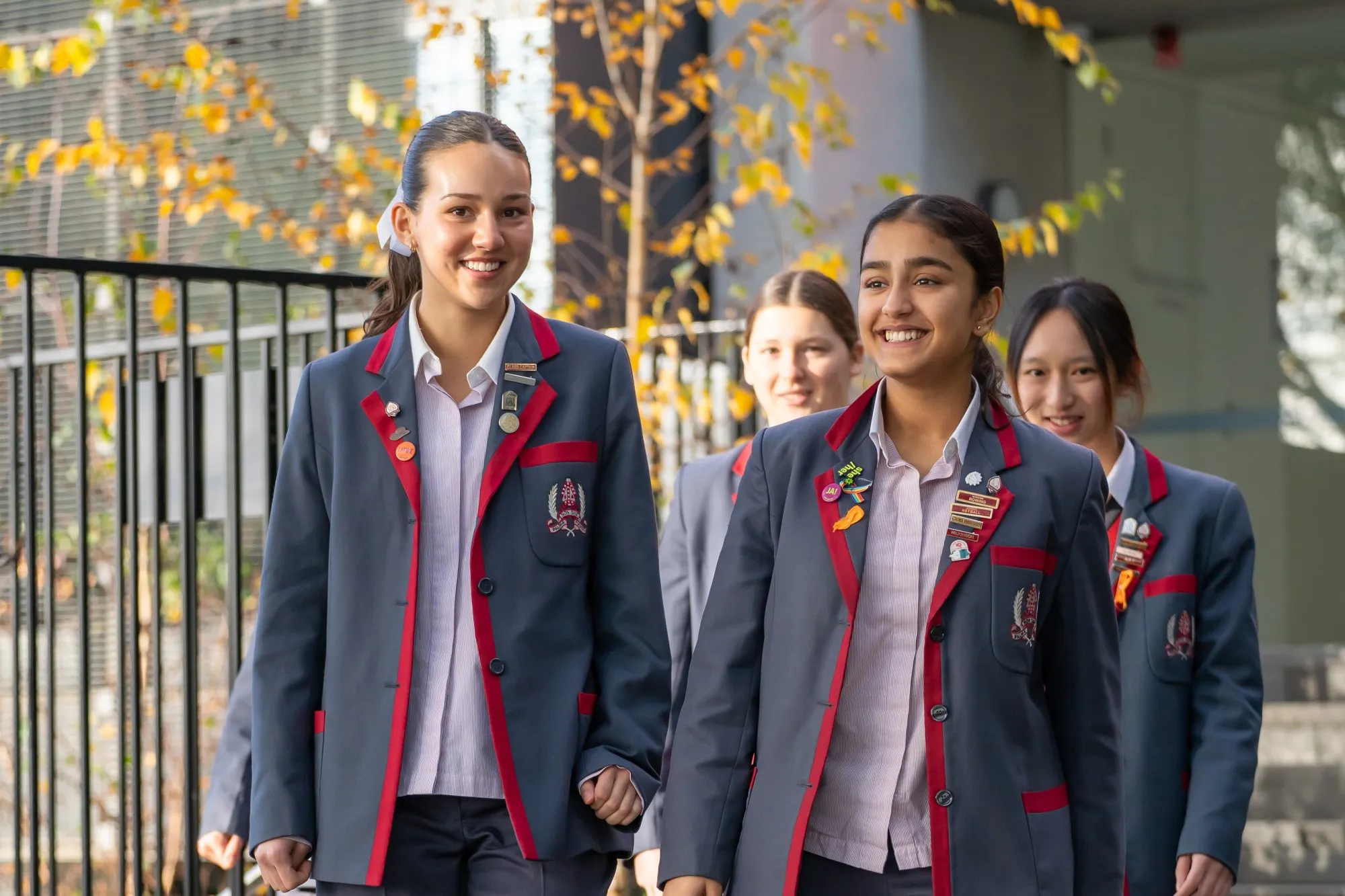 Four smiling female students wearing gray blazers with red trim and school badges walking outdoors.