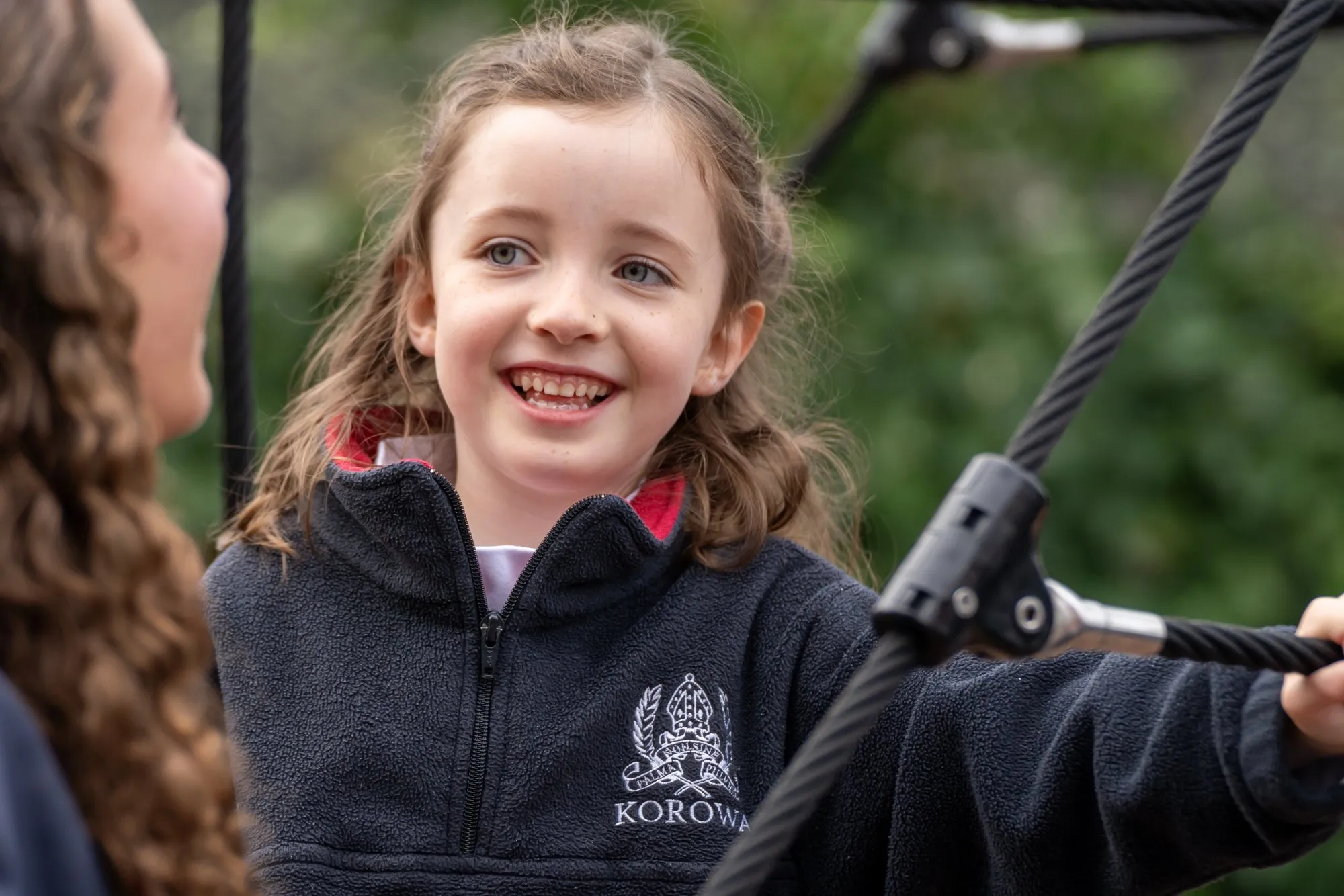 Smiling young girl in a Korowa school fleece jacket holding onto climbing ropes outdoors.