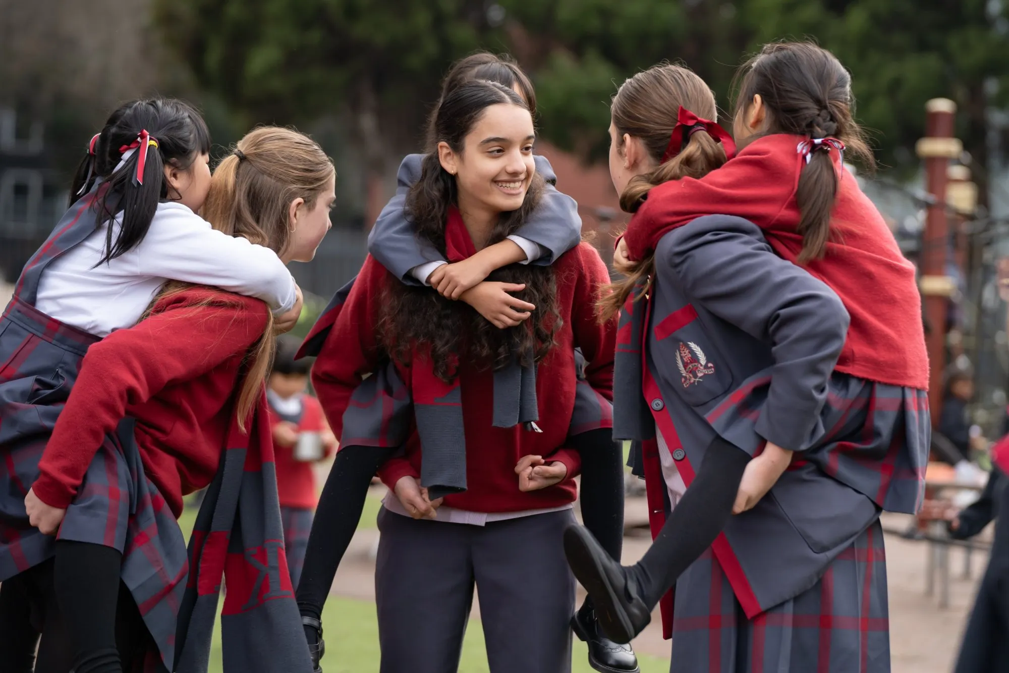 Group of schoolgirls in uniforms giving piggyback rides to each other outdoors, smiling and enjoying.
